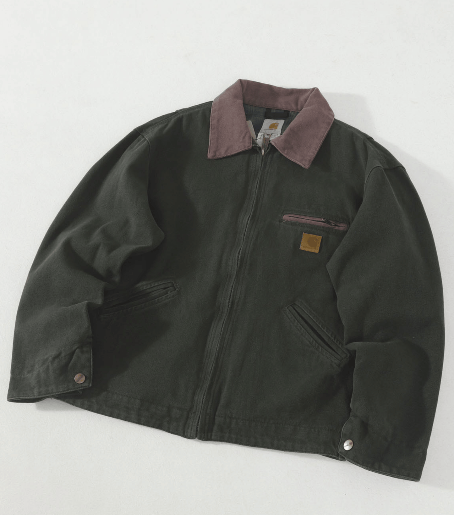 A dark olive green J97 Carhartt work jacket with a muted brown collar, featuring a front zipper, chest zip pocket, and a small brown leather Carhartt logo patch on the left chest, laid flat on a light background.