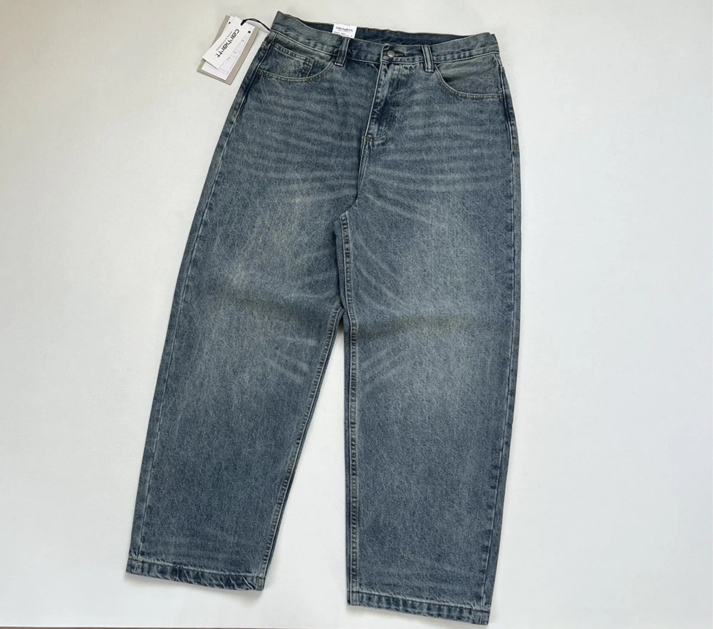 A pair of medium-wash baggy vintage Carhartt blue denim jeans laid flat on a light gray background, featuring a straight-leg fit and a white paper tag attached to the waistband.
