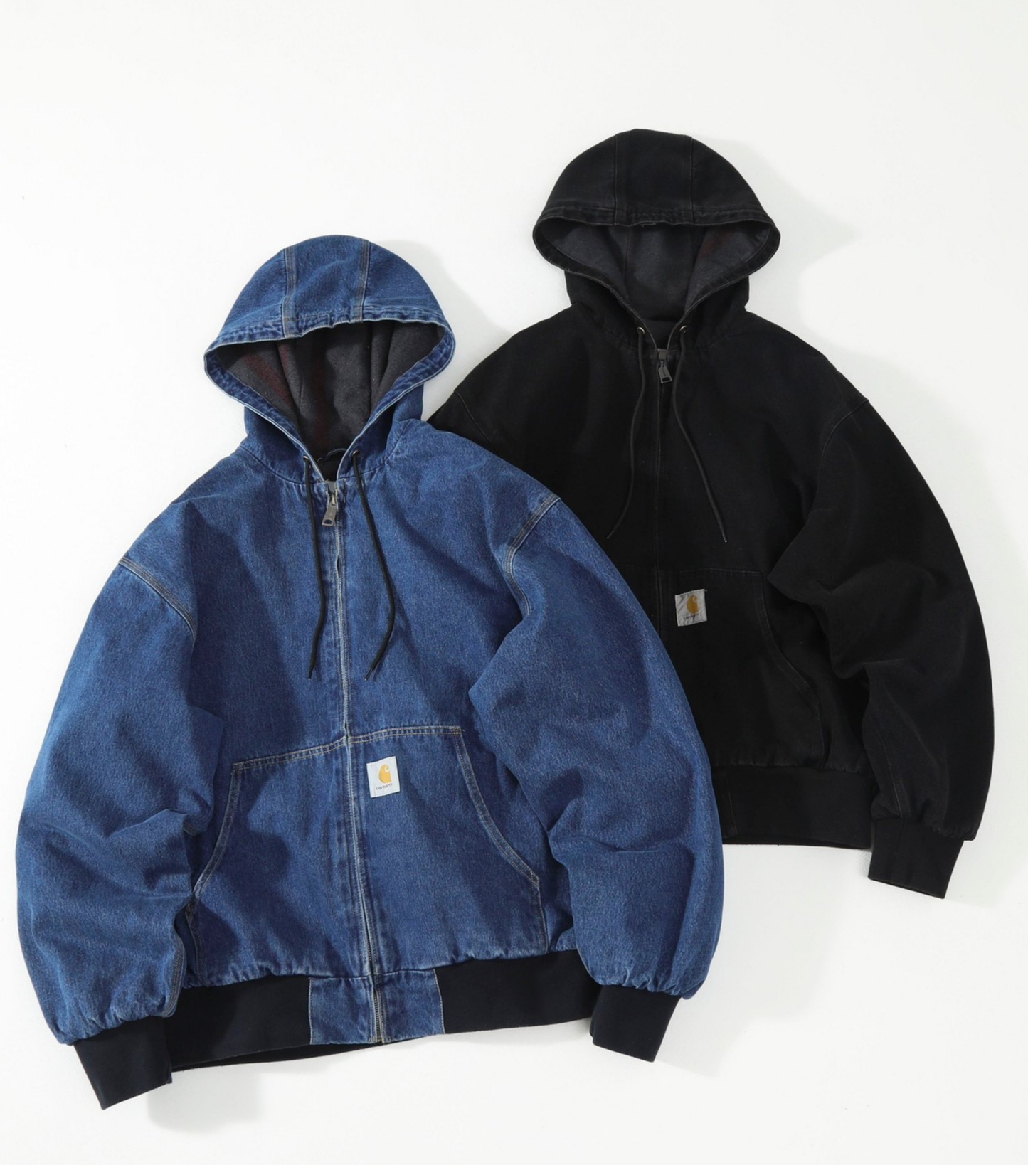 Two hooded zip-up Carhartt J140 jackets laid flat on a white background: one blue denim jacket in front and one black jacket behind it. Each jacket has a drawstring hood, front zipper, ribbed cuffs and hem, and a small rectangular Carhartt patch on the chest.