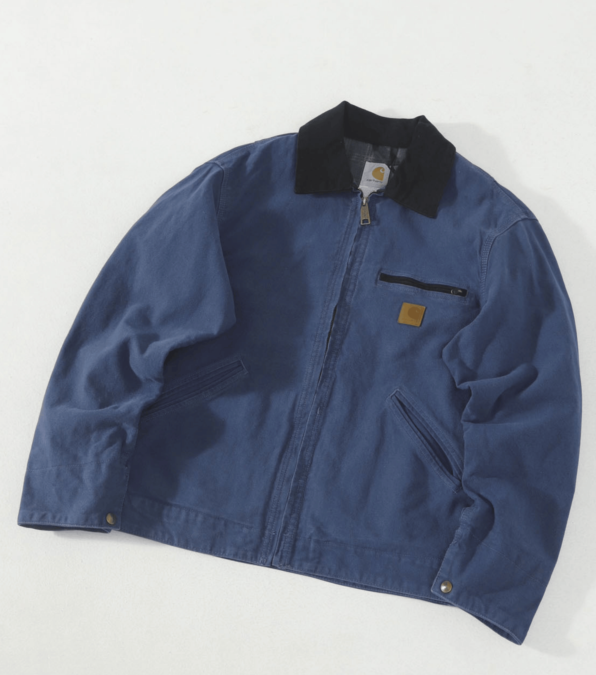 A blue Carhartt work jacket with a black collar and a small brown brand patch on the chest, laid flat on a light background.