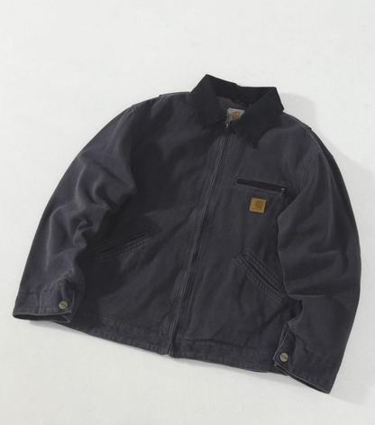 A dark charcoal gray Carhartt work jacket with a zippered front, collar, and a signature brown leather logo patch on the chest pocket, laid flat on a light gray background.