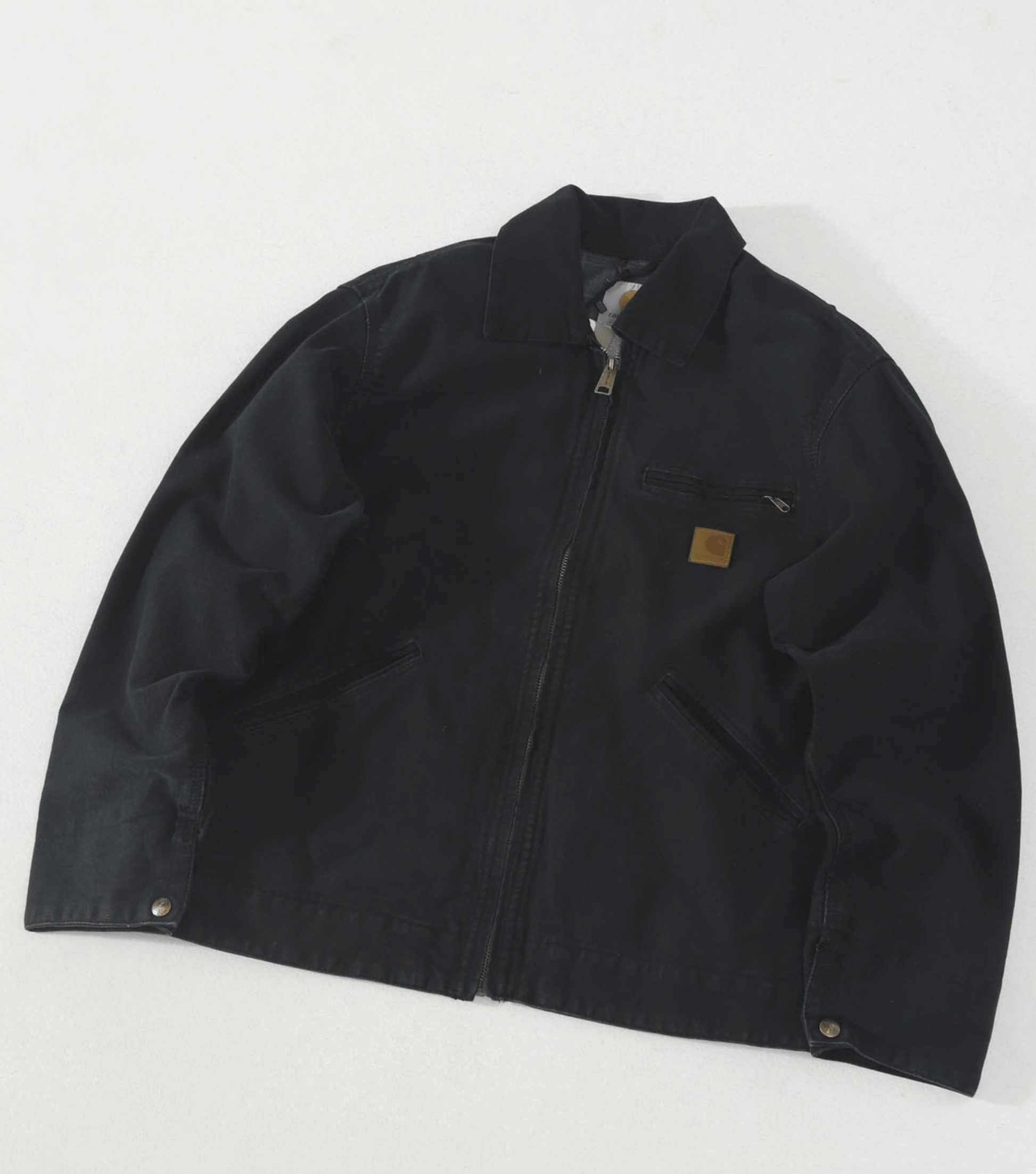 A black Carhartt J97 canvas utility jacket with a front zipper, collar, and a small brown leather Carhartt logo patch on the chest, laid flat on a white background.
