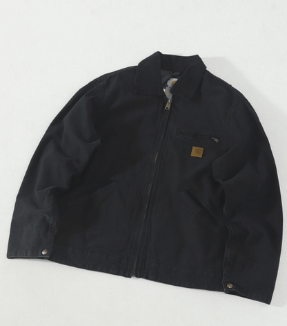 A black Carhartt J97 canvas utility jacket with a front zipper, collar, and a small brown leather Carhartt logo patch on the chest, laid flat on a white background.