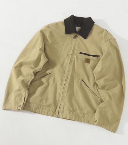 A tan Carhartt J97 vintage canvas utility jacket with a contrasting black collar, featuring a front zipper, zippered chest pocket, and zippered hand pockets.