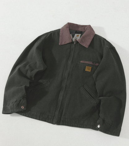 A dark olive green J97 Carhartt work jacket with a muted brown collar, featuring a front zipper, chest zip pocket, and a small brown leather Carhartt logo patch on the left chest, laid flat on a light background.