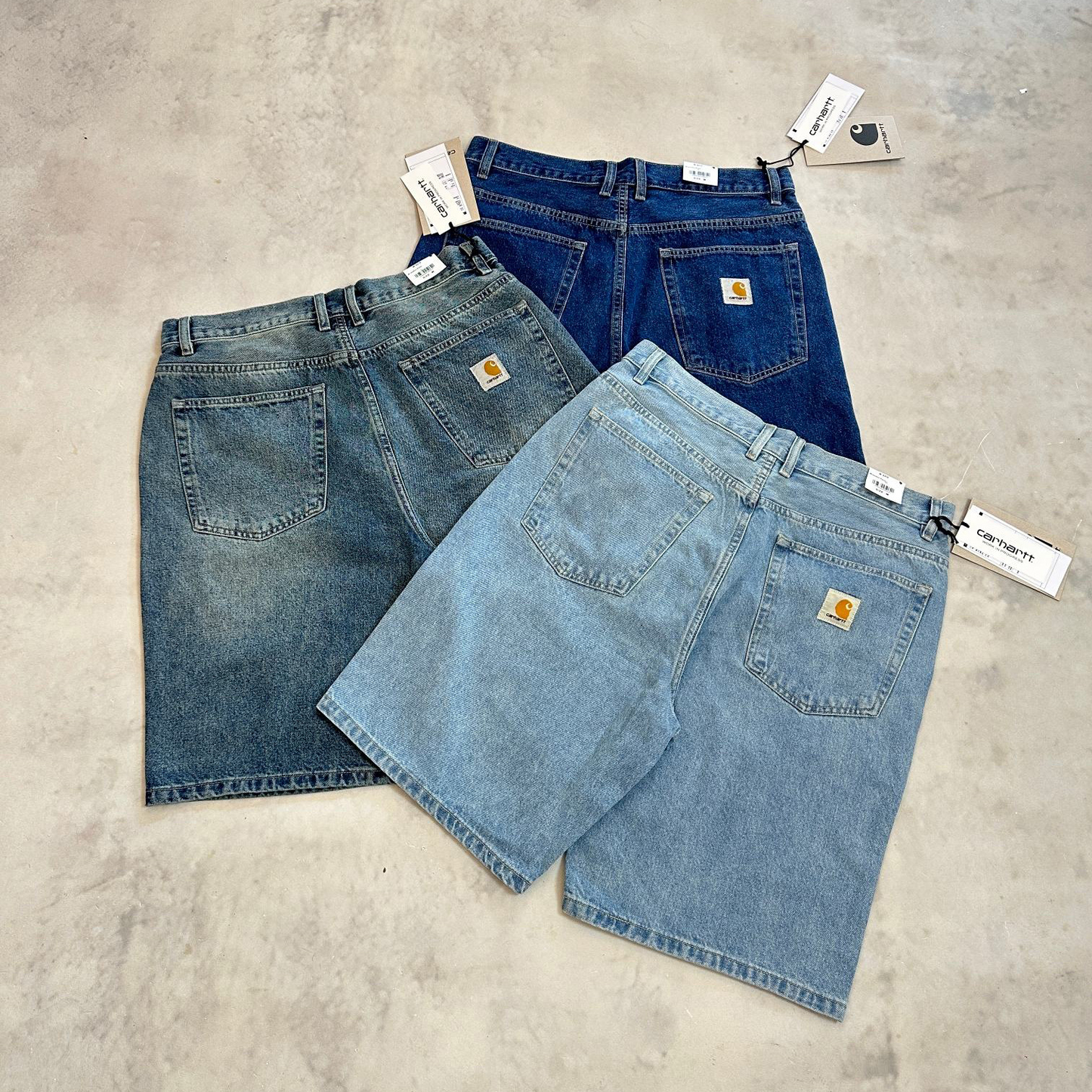 Three pairs of Carhartt denim shorts in different washes (light blue, medium blue, and dark blue) are arranged on a textured concrete floor, showing the back pockets and Carhartt brand patches.