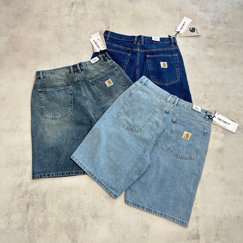Three pairs of Carhartt denim shorts in different washes (light blue, medium blue, and dark blue) are arranged on a textured concrete floor, showing the back pockets and Carhartt brand patches.