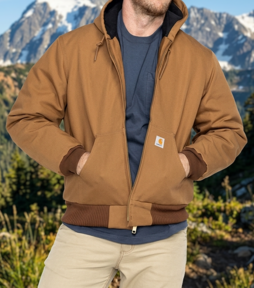 A man wearing a brown J140 Carhartt hooded jacket over a navy blue t-shirt and khaki pants, standing outdoors with a mountain background.