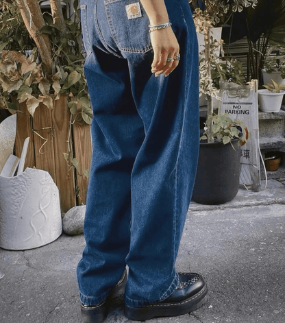 A person wearing very loose, wide-leg blue carhartt denim jeans with black platform shoes, standing outdoors on a paved surface near potted plants and a wooden fence.