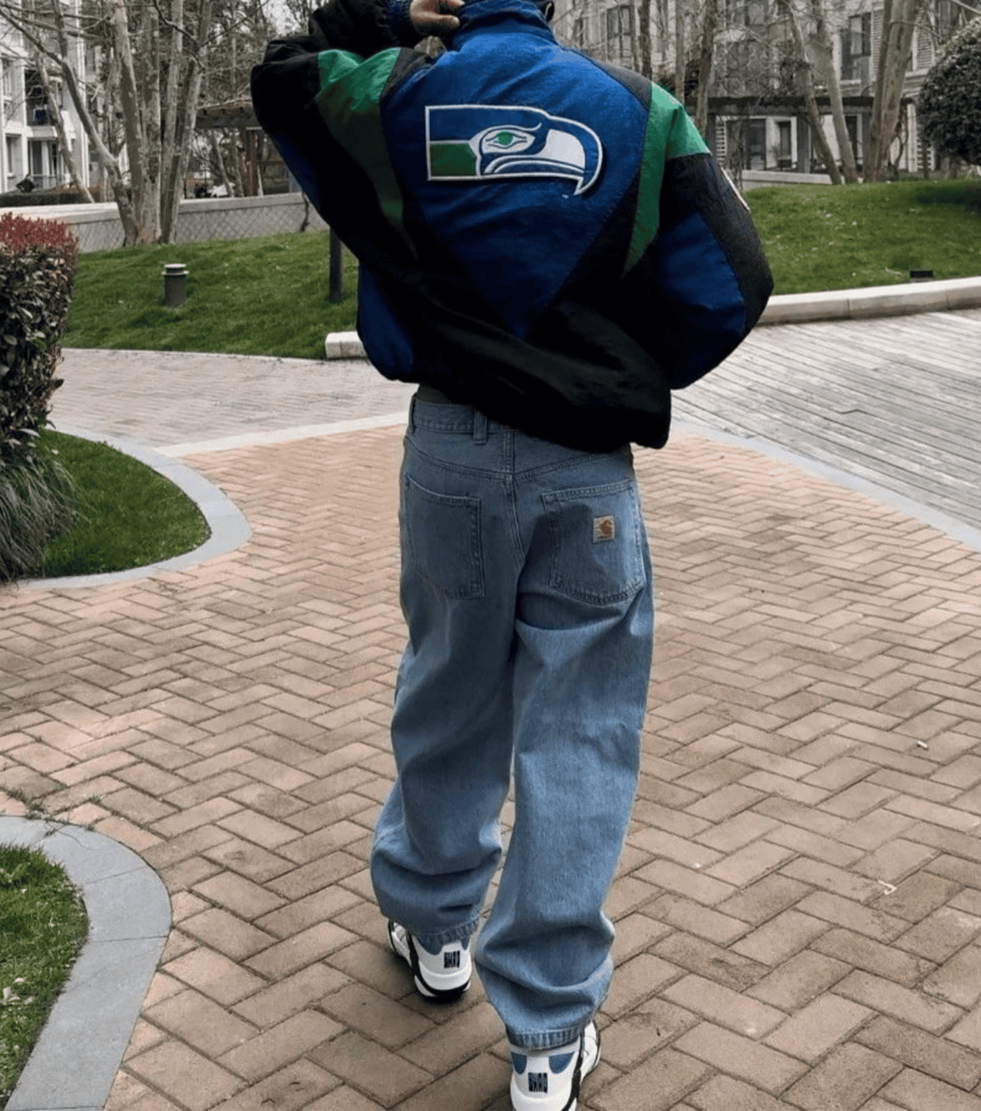 A person viewed from behind wearing a blue and green Seattle Seahawks varsity jacket, light blue baggy jeans, and white sneakers, standing on a brick walkway in an outdoor park setting.