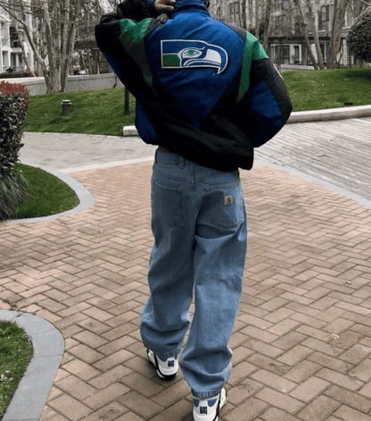 A person viewed from behind wearing a blue and green Seattle Seahawks varsity jacket, light blue baggy jeans, and white sneakers, standing on a brick walkway in an outdoor park setting.