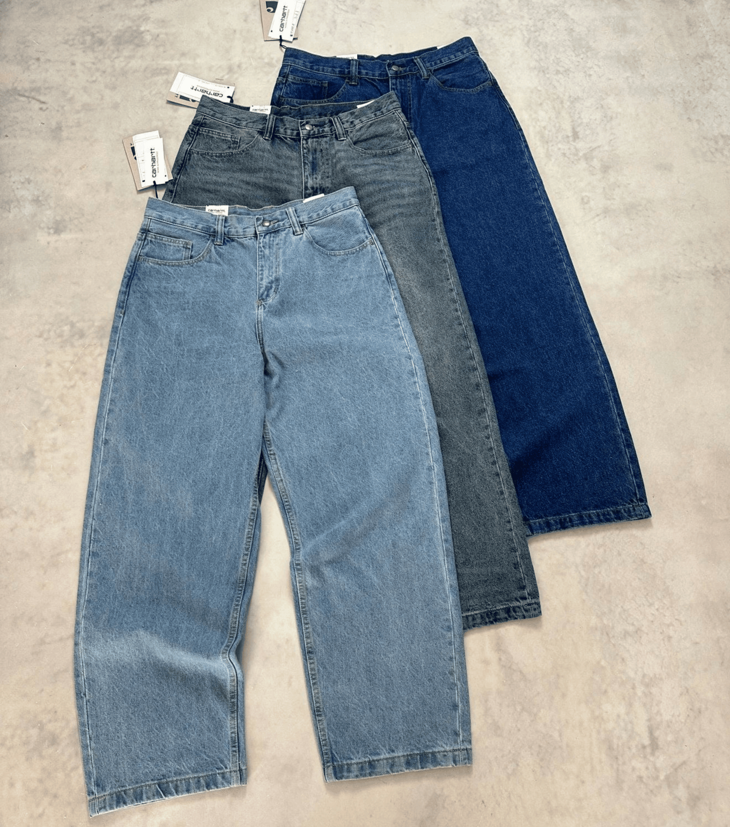 Three pairs of wide-leg Carhartt vintage jeans in light blue, medium gray, and dark blue denim, arranged diagonally on a textured concrete floor with tags attached.