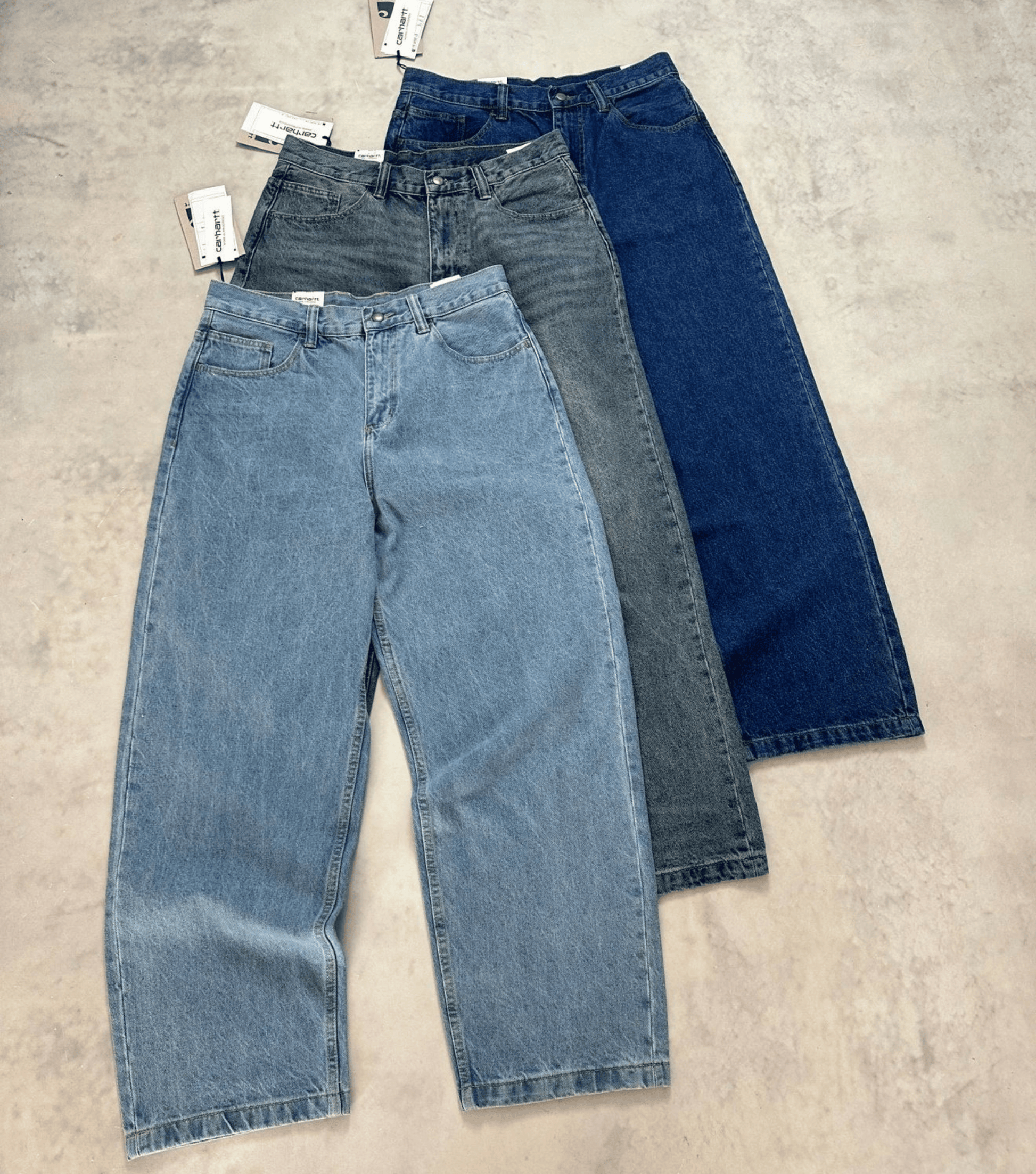 Three pairs of wide-leg Carhartt vintage jeans in light blue, medium gray, and dark blue denim, arranged diagonally on a textured concrete floor with tags attached.