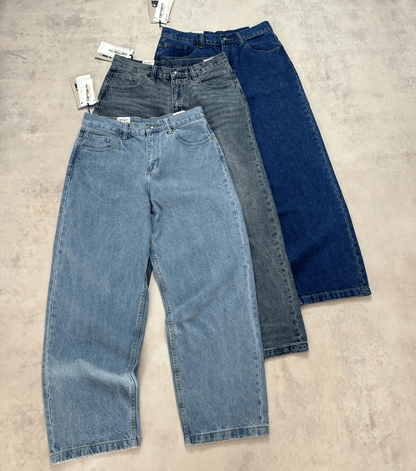Three pairs of wide-leg Carhartt vintage jeans in light blue, medium gray, and dark blue denim, arranged diagonally on a textured concrete floor with tags attached.