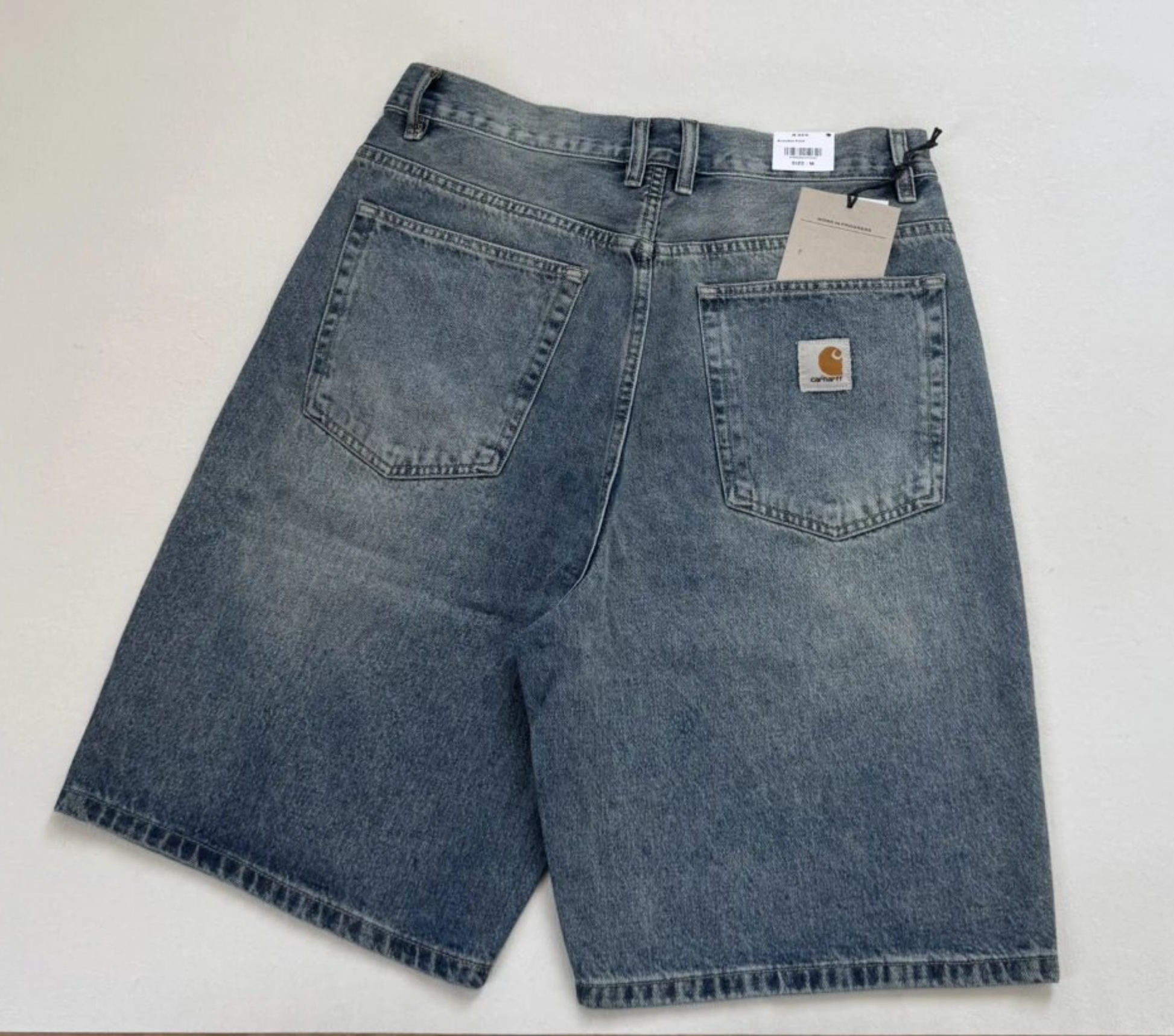 Carhartt denim shorts in a faded medium blue wash, shown from the back with the brand's signature logo patch on the pocket and retail tags attached.