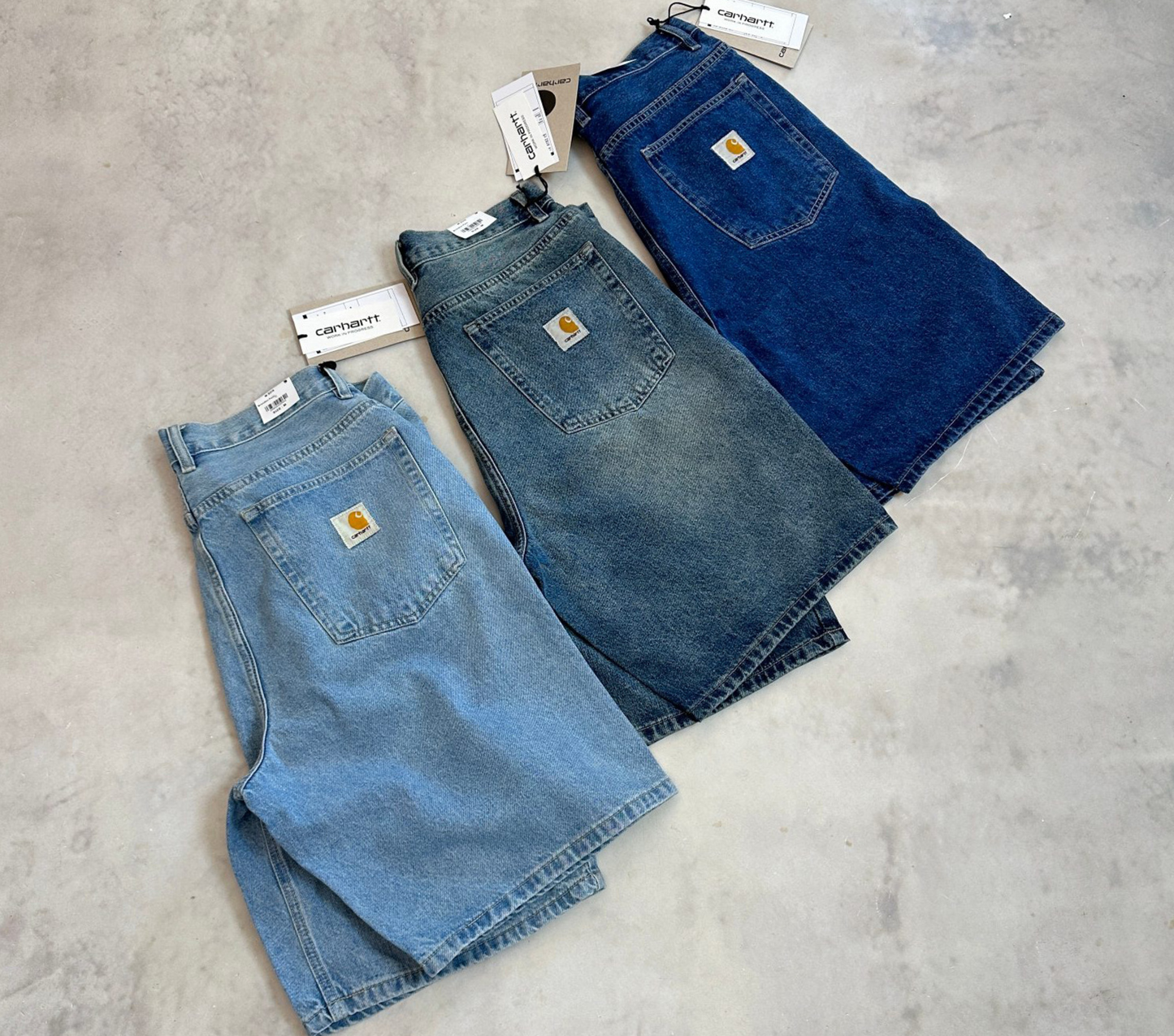 Three pairs of Carhartt denim shorts in light blue, medium blue, and dark blue washes, arranged diagonally on a light gray concrete floor. Each pair has its original white Carhartt tags attached.