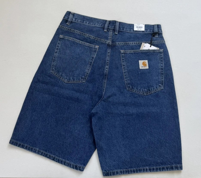 A pair of blue Carhartt denim shorts shown from the back, featuring a classic five-pocket design and a visible Carhartt logo patch on the right pocket, laid flat on a light background.