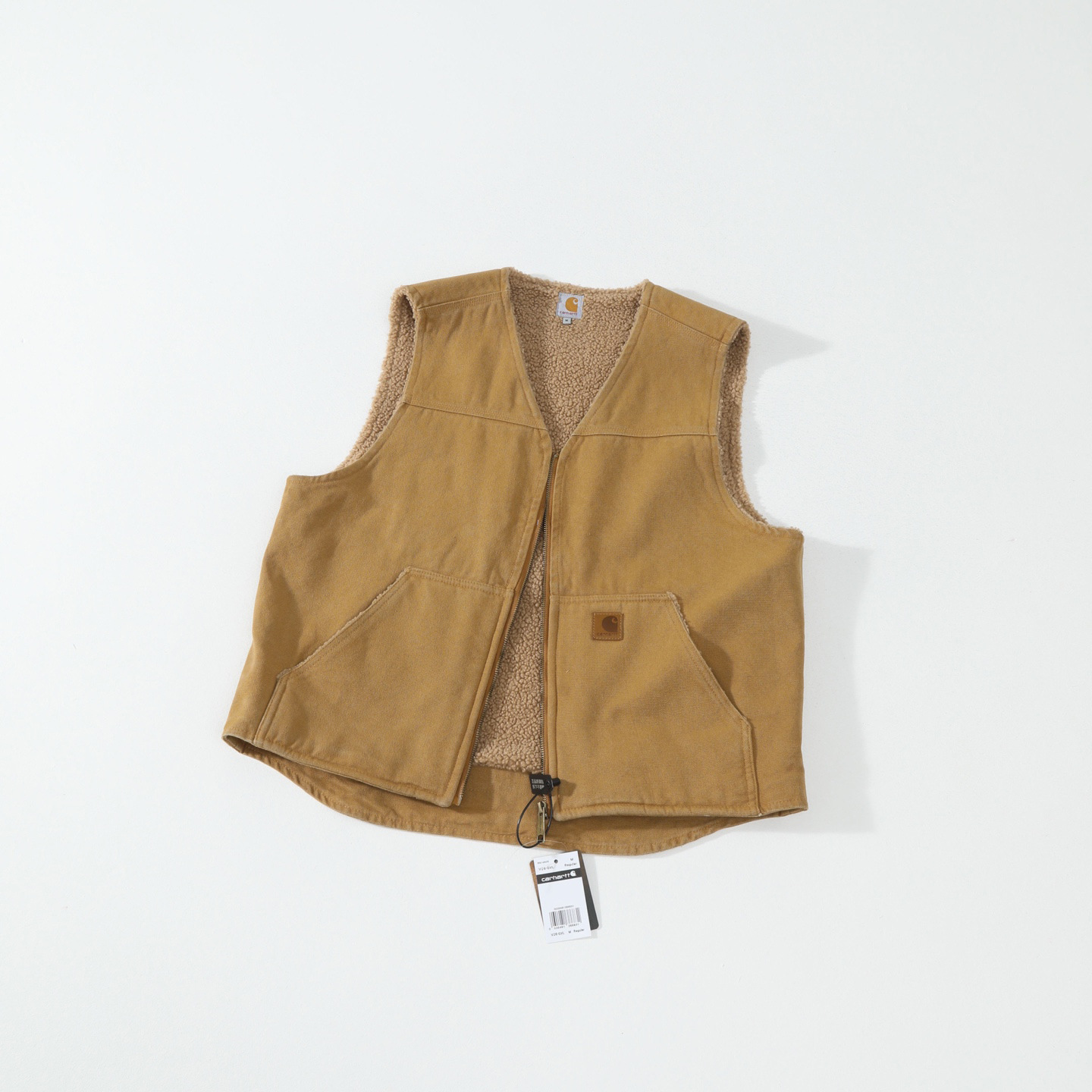 A tan corduroy carhartt vest with a brown sherpa lining, laid flat on a white background. It features a small brown leather patch on the front pocket
