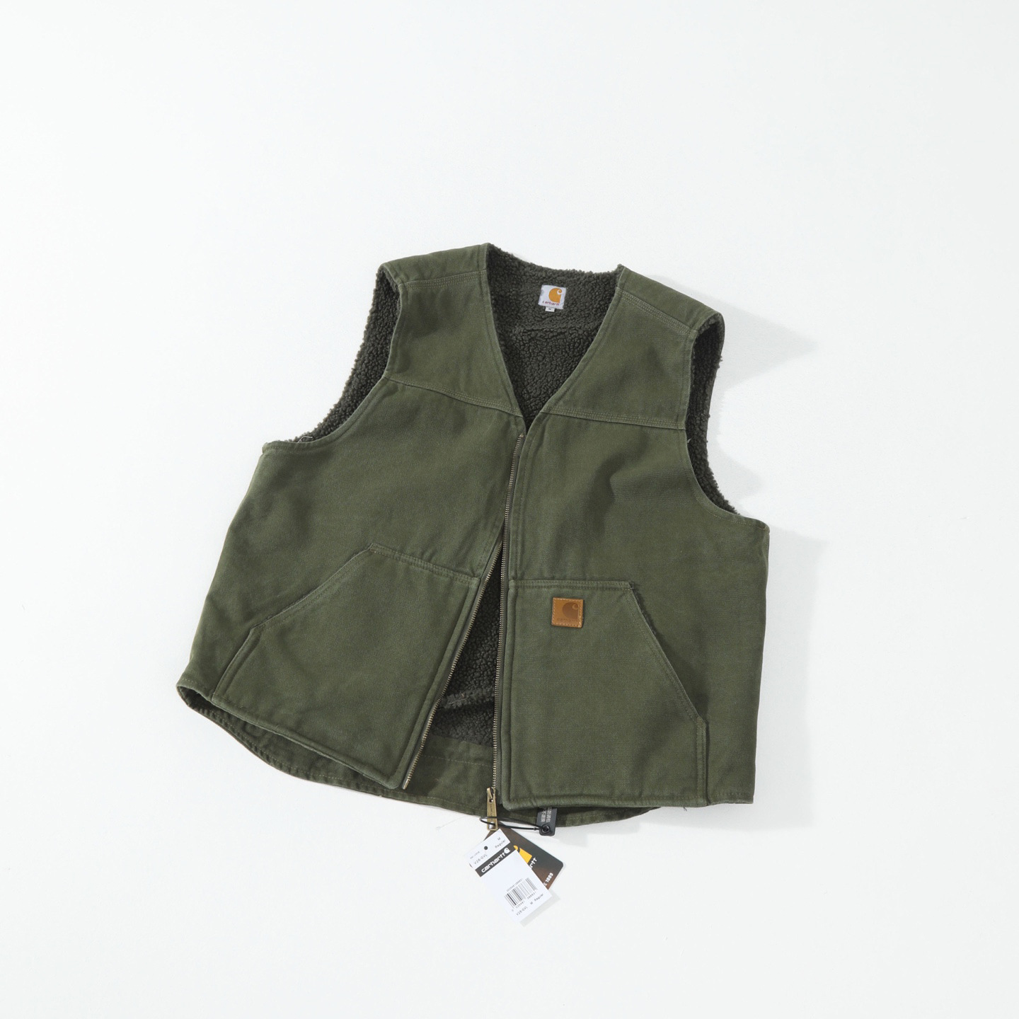 An olive green Carhartt vest with a black sherpa lining, laid flat on a white background with a small leather logo patch and hangtag visible.